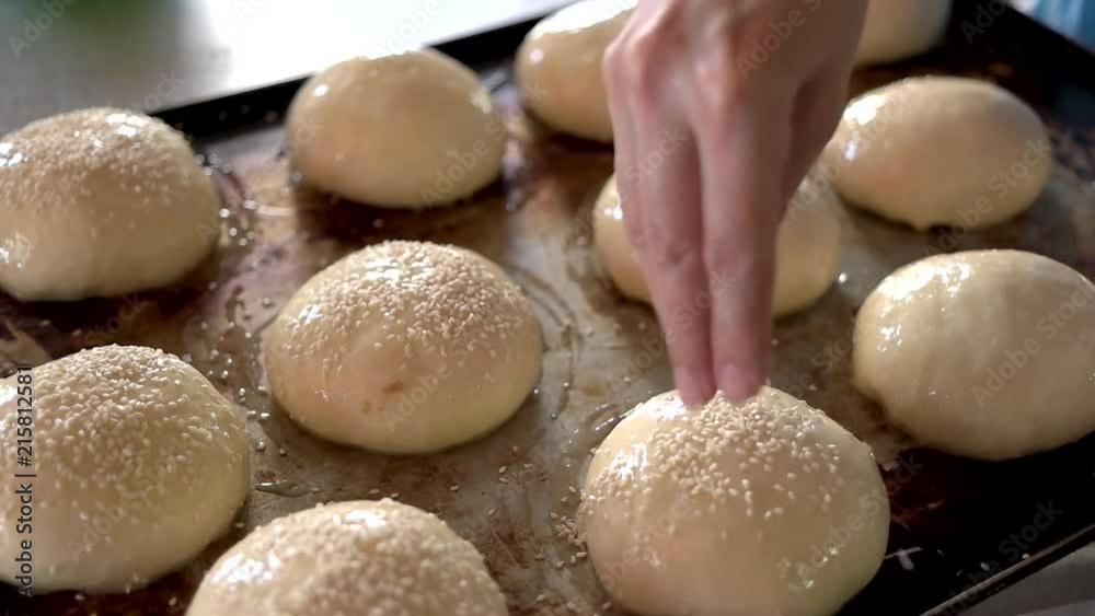 Hand spreading sesame seeds on dough. Unbaked buns on oven tray. Baking of traditional french buns.
