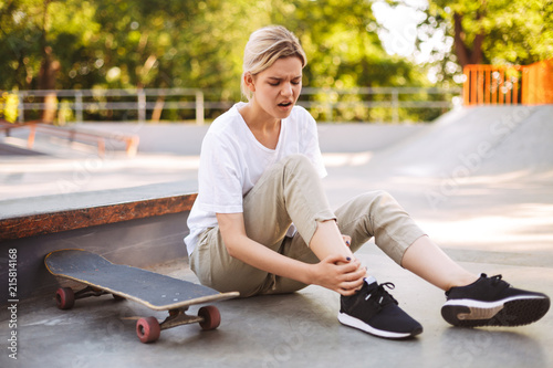 Upset skater girl holding her painful leg with skateboard near while spending time at skatepark