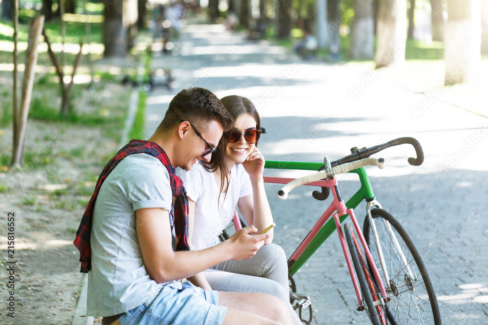 Obraz premium Young romantic couple with bicycle sitting on beanch and speaking in the sunny summer park