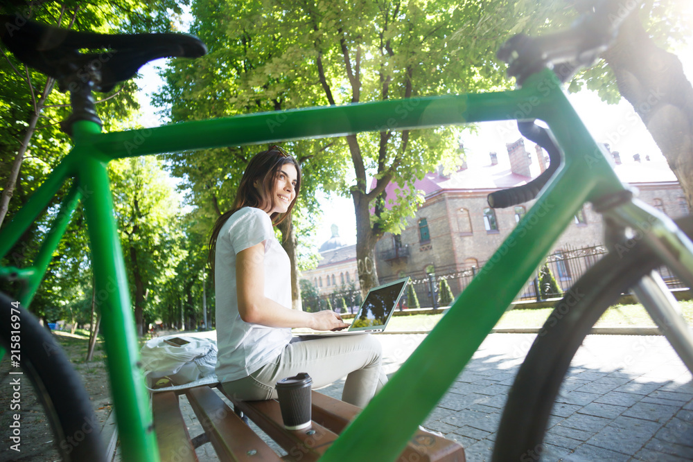 Pretty girl with green bicycle sitting on beanch and working with laptop in the sunny summer park, she drinking coffee and thinking about something