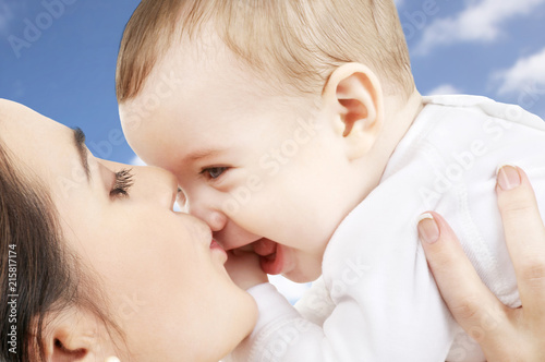 family and motherhood concept - happy smiling young mother kissing little baby over sky background