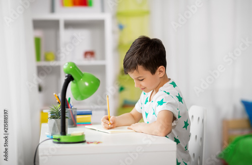 education, childhood and school concept - little boy sitting at desk and writing to notebook at home