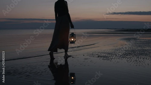 Girl in dress walking on beach and holding old kerosene lantern in her hand after sunset at night