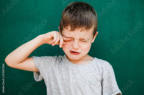 A little boy on a green background wipes his tears with his hands.
