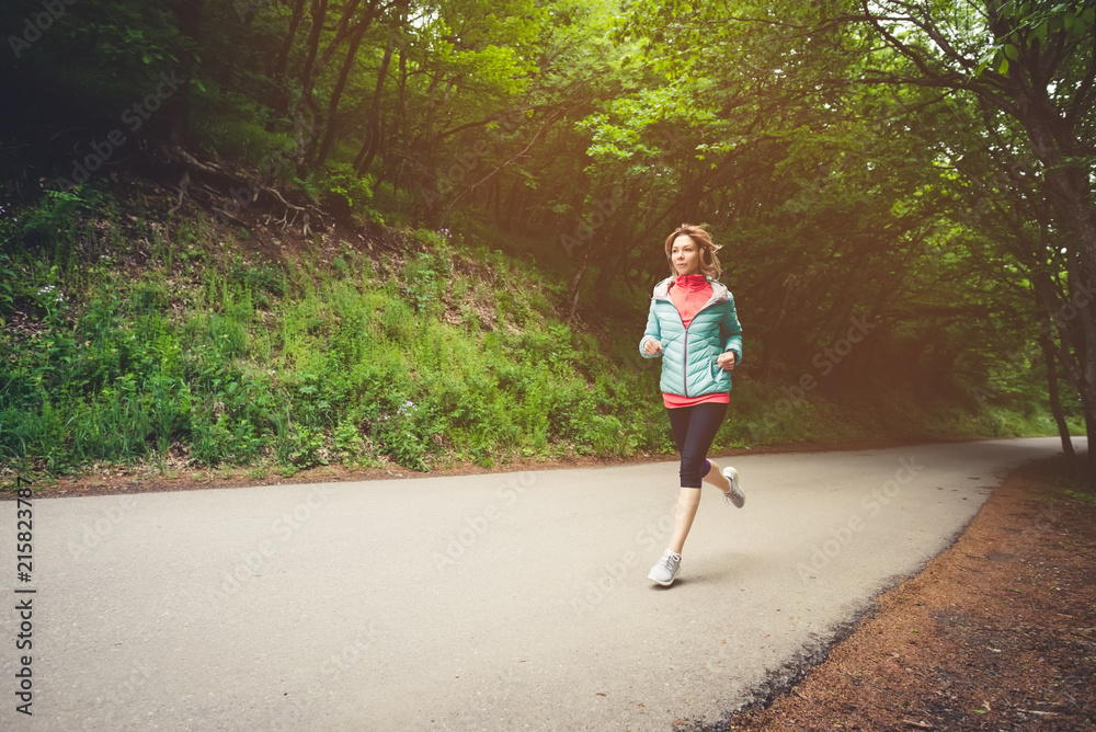 Young fitness blonde woman in headphones running at morning caucasian forest trail in sun light Young fitness blonde woman in headphones running at morning caucasian forest trail in sun light