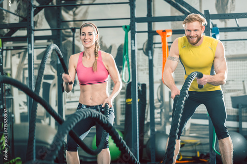 Low-angle view of a strong and competitive couple smiling while exercising together with battle ropes during functional training at the gym