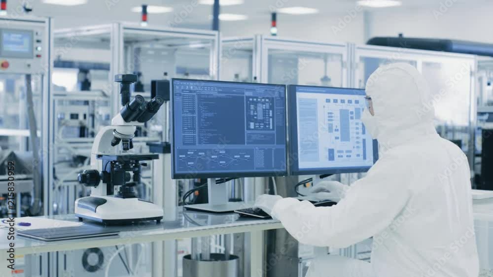In Laboratory Over the Shoulder View of Scientist in Protective Clothes Doing Research on a Personal Computer. Modern Manufactory Producing Semiconductors and Pharmaceutical Items.