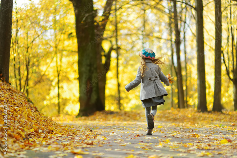 Obraz premium Cute little girl having fun on beautiful autumn day. Happy child playing in autumn park. Kid gathering yellow fall foliage.