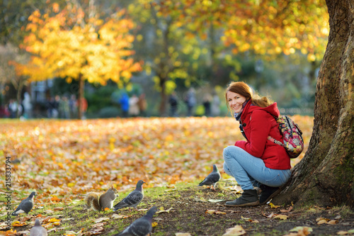 Young female tourist feeding squirrels and pigeons in St James's Park in London, United Kingdom, on beautiful sunny autumn day