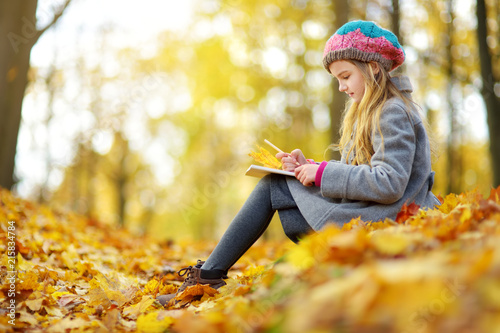 Cute little girl sketching outside on beautiful autumn day. Happy child playing in autumn park. Kid drawing with colourful pencils.