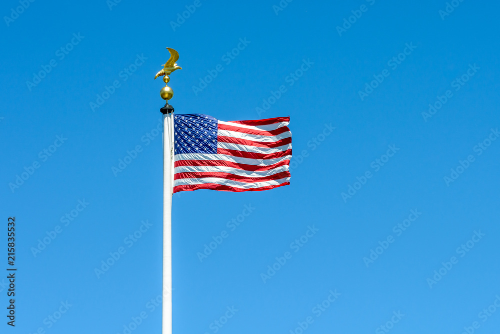 The flag of the United States of America blowing in the wind at full-mast on a white pole topped with a golden eagle on ball against blue sky.