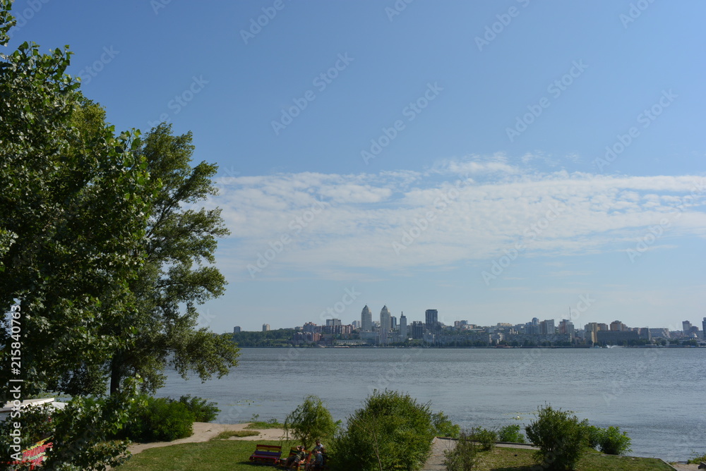 Сity landscape with green trees on the left bank of the city, river and new houses on the second shore and white clouds above them Сity landscape with green trees on the left bank of the city, river and new houses on the second shore and white clouds above them