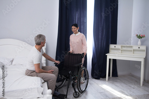 Nurse bringing wheelchair to patient. Beautiful asian caregiver pushing wheelchair to bed on which elderly man is sitting.