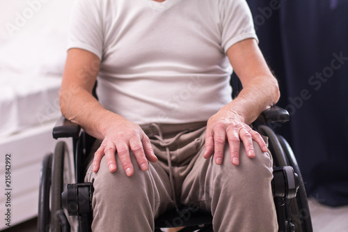 Elderly man sitting in wheelchair. Senior man wearing gray shirt and beige trousers sitting in wheelchair with his hands on his knees.