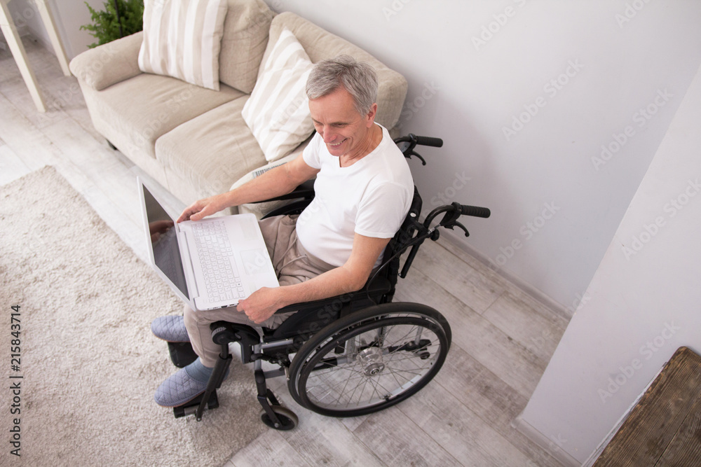 Obraz premium Disabled aged man sitting in wheelchair using laptop. Top view of old man smiling while sitting in his living room in wheelchair and websearching via computer.
