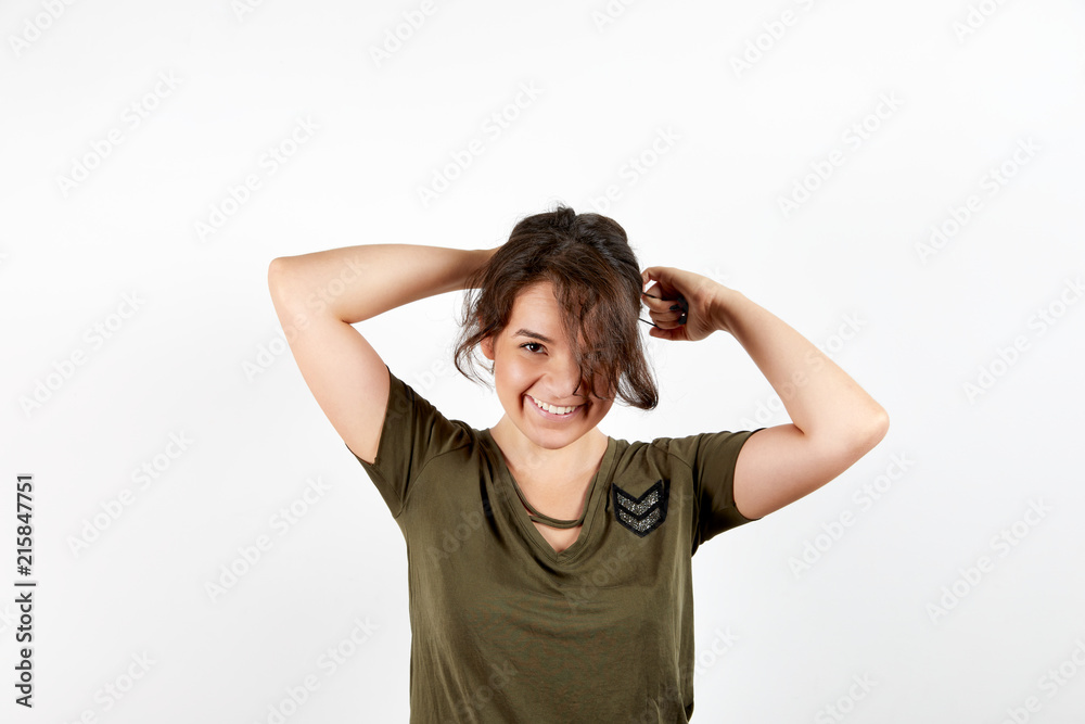 Adorable young tanned female in dark green shirt smiling and looking at camera doing ponytail standing over white studio background Adorable young tanned female in dark green shirt smiling and looking at camera doing ponytail standing over white studio background