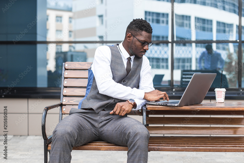 african businessman in suit and glasses is sitting at a table with a Laptop, a cup of coffee against a background of glass buildings african businessman in suit and glasses is sitting at a table with a Laptop, a cup of coffee against a background of glass buildings