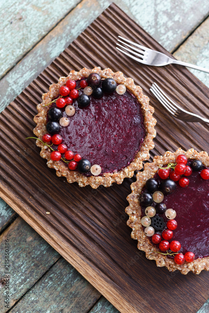 Small vegan tarts made of nuts and berry jam decorated with black, red and white currants on oak cutting board Small vegan tarts made of nuts and berry jam decorated with black, red and white currants on oak cutting board
