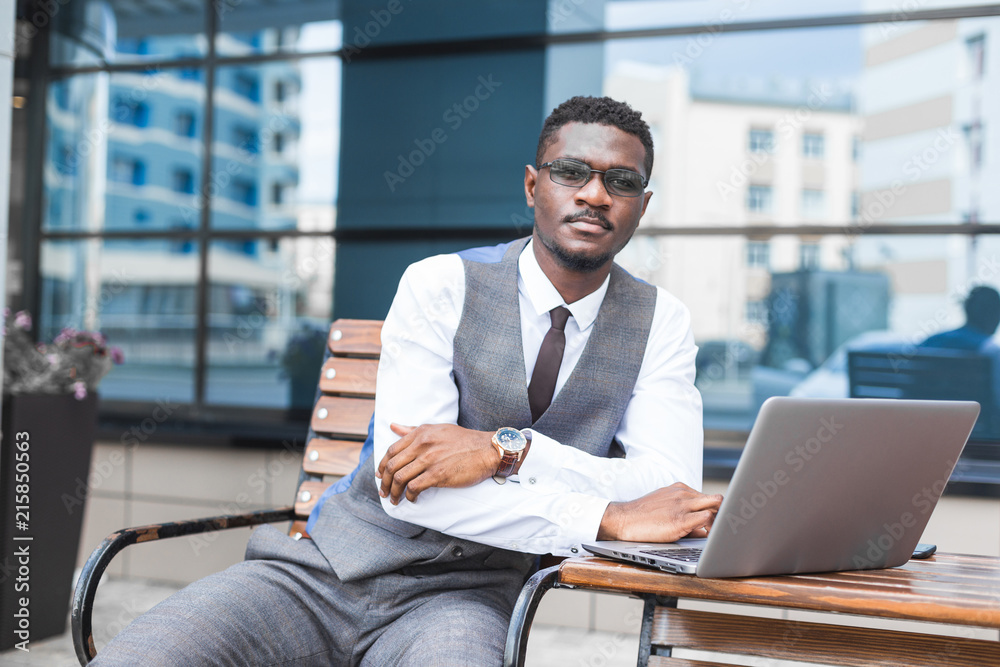 african businessman in suit and glasses is sitting at a table with a Laptop, a cup of coffee against a background of glass buildings