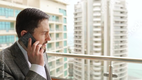 Businessman angrily talking on phone, standing on balcony. Big city background