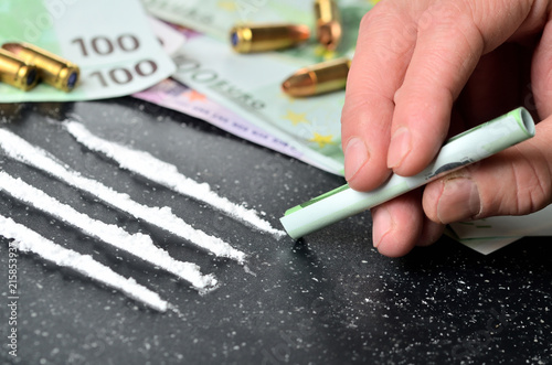 Hand of man snorting cocaine, euro banknotes and gun ammunition in background