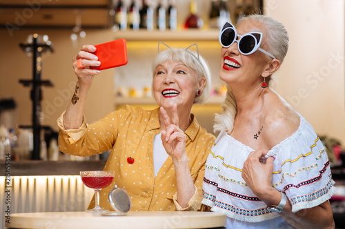 Happy moments. Delighted aged woman showing V sign while taking a selfie together with her friend