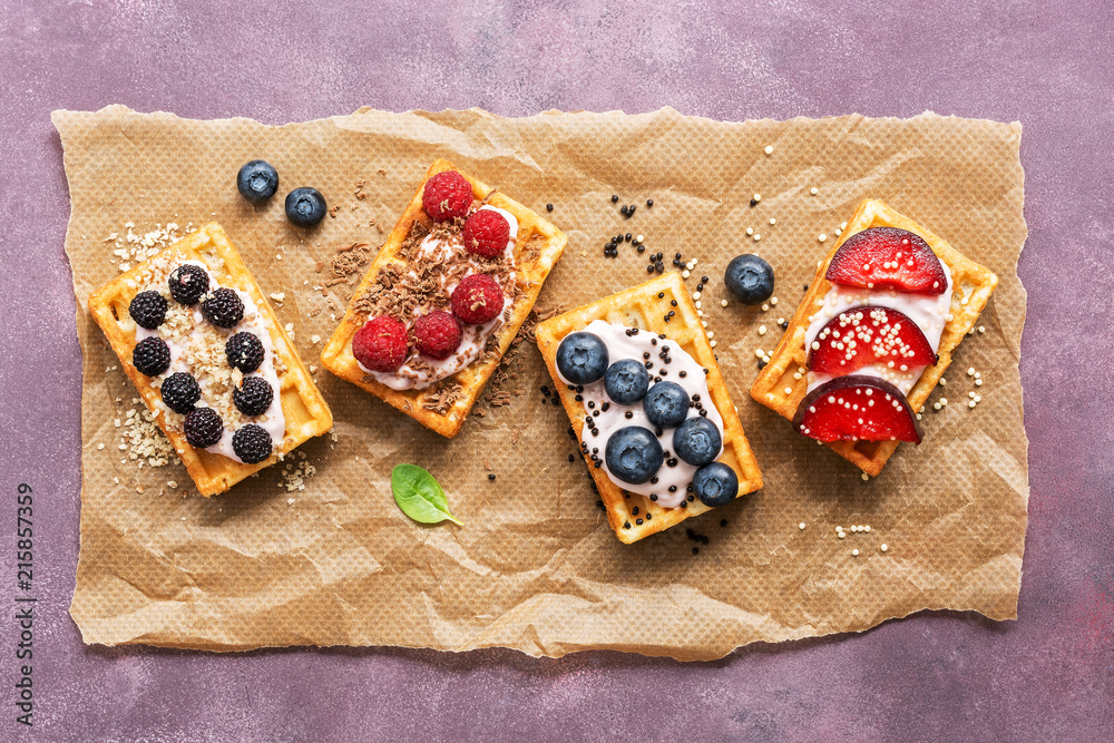 Homemade waffles with berries and fruits, blueberries, peaches, raspberries, black raspberries on a purple background. Waffles on parchment.Top view, flat lay. Homemade waffles with berries and fruits, blueberries, peaches, raspberries, black raspberries on a purple background. Waffles on parchment.Top view, flat lay.