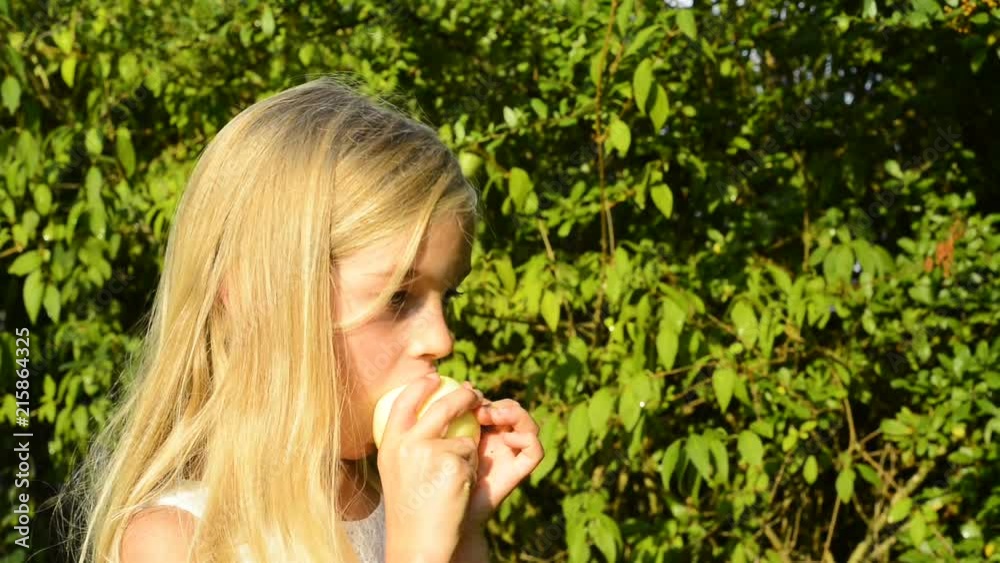 Child cute girl eating apple outside in garden during sunset. Greenery background Child cute girl eating apple outside in garden during sunset. Greenery background