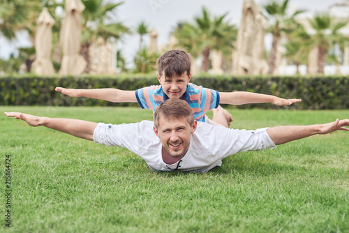 Father and son laying at the green grass at the hotel territory. They are having fun, smiling and looking to the camera. Their hand sare open wide. They look like planes.