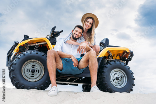 bottom view of happy young couple sitting on ATV on sandy dune in front of cloudy sky