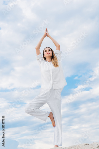 fit young woman practicing yoga in tree pose while standing on sand dune