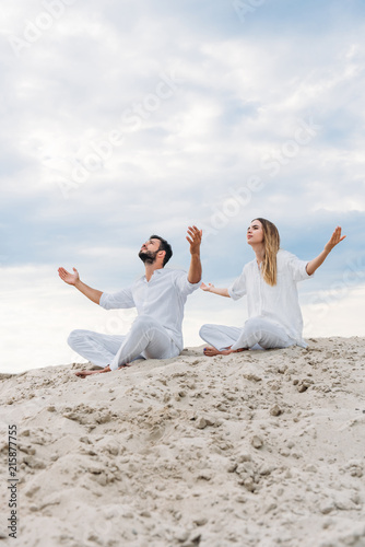 young fit couple on yogi meditating while sitting on sandy dune in lotus pose (padmasana)