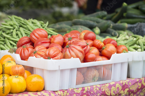 White plastic container of red heirloom tomatoes on a farmer's market table.  Green peas, okra and zucchini in the background.  Yellow tomatoes in the foreground.