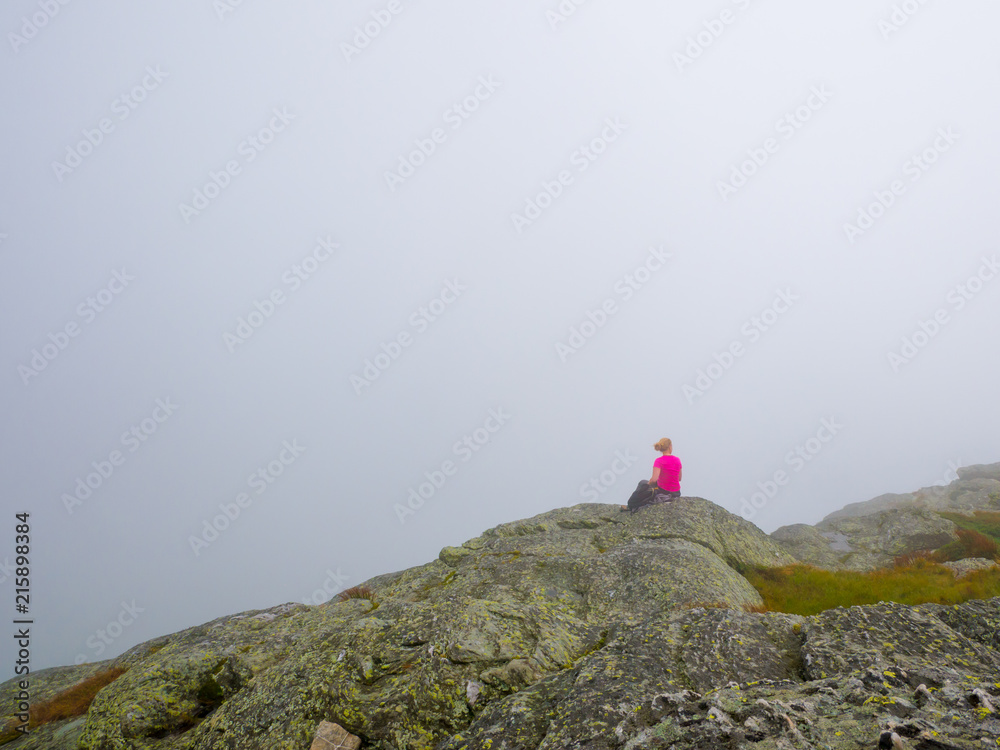 A lonely woman sitting on the top of a mountain looking at the horizon on a foggy summer day