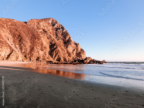 Shore of the Pacific Ocean at Pfeiffer Beach near Big Sur, California. Evening coastal scene with clear blue sky and rocks colored red by the sunset. Some reflections are visible in the wet sand.