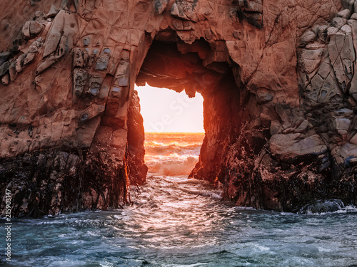 Stunning view of the Pacific Ocean through an opening in the rocky cliff at Pfeiffer Beach, near Big Sur, California. Rocks colored red by the sunset and covered with algae. Glowing gates to the sea.