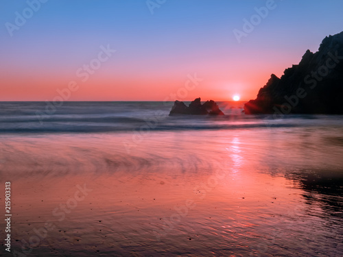 Long-exposure photography of jagged coastal rocks on the Pacific Coast at sunset. A low angle shot taken at Pfeiffer Big Sur Beach, California. Soft sun starburst on the horizon between two rocks.