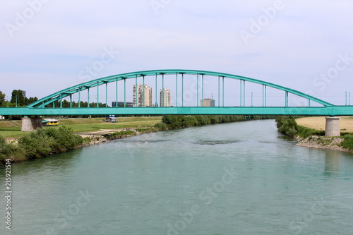 Old green metal arched railway bridge on two strong concrete pillars covered in stone over mighty river with apartment buildings in background © hecos
