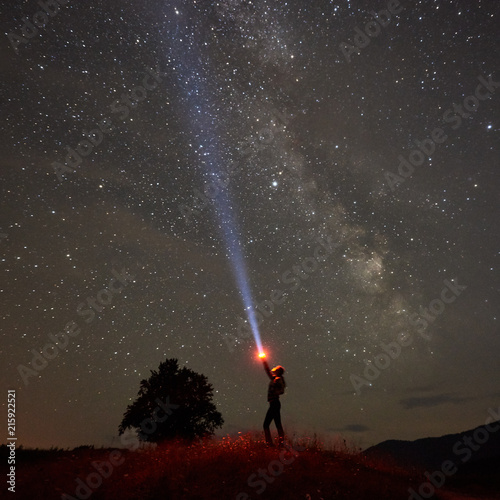 Silhouette of woman standing against night starry sky with Milky Way in the mountains with a flashlight in his hand