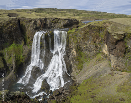 View on Beautiful Fragifoss waterfall on Geirlandsa river in South Iceland, green moss and rock, blue sky background