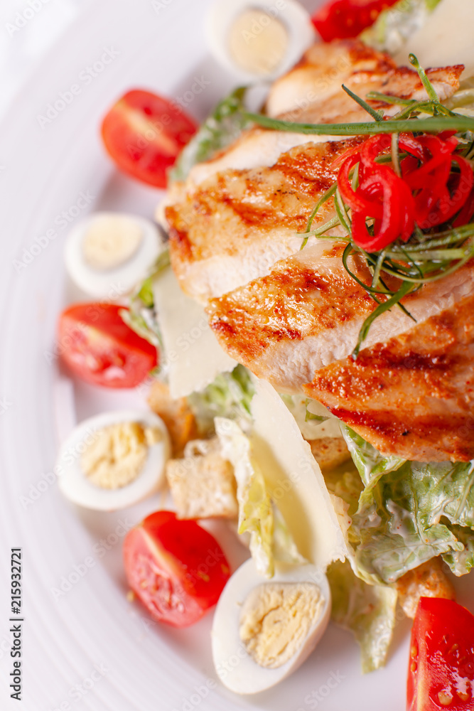 Caesar salad with quail eggs, cherry tomatoes, Parmesan cheese and grilled chicken in white plate on light table.