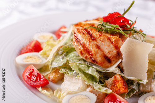 Caesar salad with quail eggs, cherry tomatoes, Parmesan cheese and grilled chicken in white plate on light table.