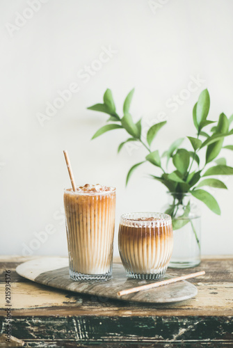 Iced coffee in tall glasses with milk and straws on board, white wall and green plant branches at background, copy space. Summer refreshing beverage ice coffee drink concept