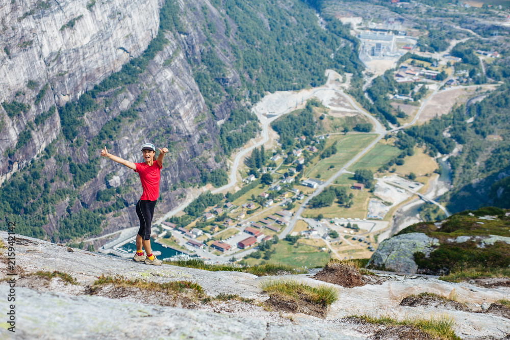 Adventure, travel, tourism, hike and people concept - cheering woman in pink t-shirt show thumbs up over mountains background. Female admiring views, relaxing during climbing on Kjeragbolten, Norway