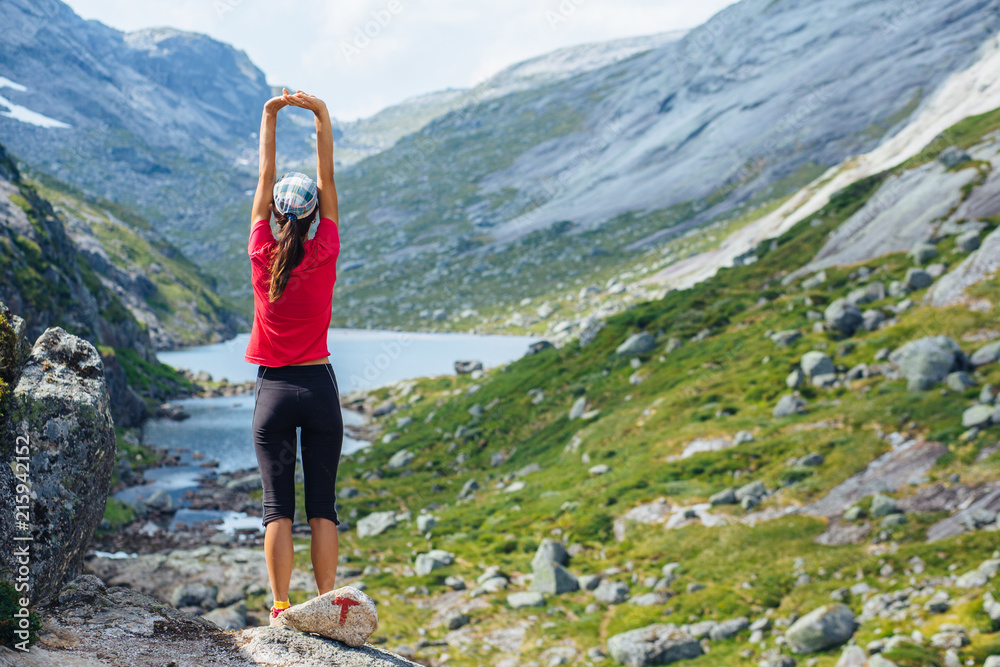 Adventure, travel, tourism, hike and people concept - slim woman in pink t-shirt stretch arms over mountains nature background. Female admiring views, relaxing during climbing on Kjeragbolten, Norway
