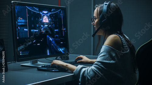 Beautiful Professional Gamer Girl Playing in First-Person Shooter Online Video Game on Her Personal Computer. Casual Cute Geek Girl Wearing Headset. In the Underground Gaming Club.