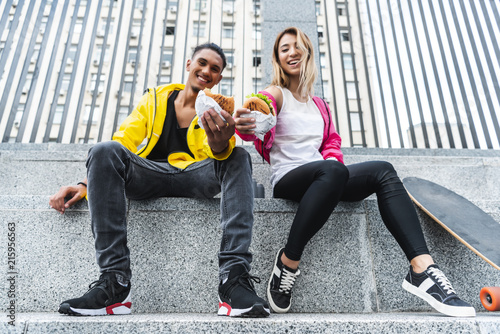 low angle view of smiling multiethnic couple of skateboarders showing burgers at city street