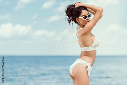 attractive brunette girl posing in sunglasses and white bikini near the sea on resort