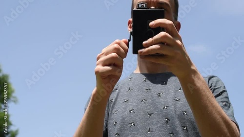 Fotógrafo con camiseta de pájaros haciendo fotos o tomando imágenes en movimiento con una cámara antigua de película con una manivela. Imagen al aire libre en el campo bajo un cielo azul despejado.