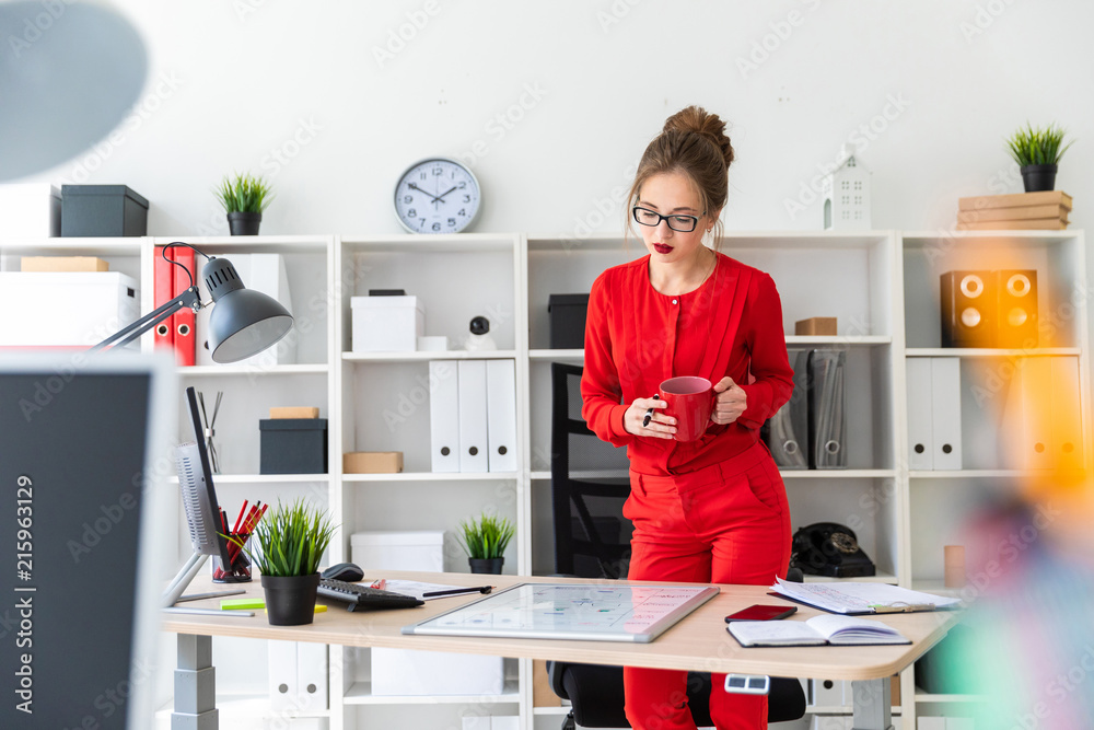 A young girl stands near a table in her office, holds a marker and a red cup in her hands and looks at the magnetic board. A young girl stands near a table in her office, holds a marker and a red cup in her hands and looks at the magnetic board.